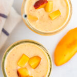 Two glass mason jars of peach smoothies. Two gold straws are in the smoothie and the smoothies are topped with chopped peached.