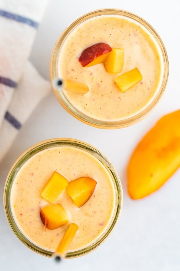 Two glass mason jars of peach smoothies. Two gold straws are in the smoothie and the smoothies are topped with chopped peached.