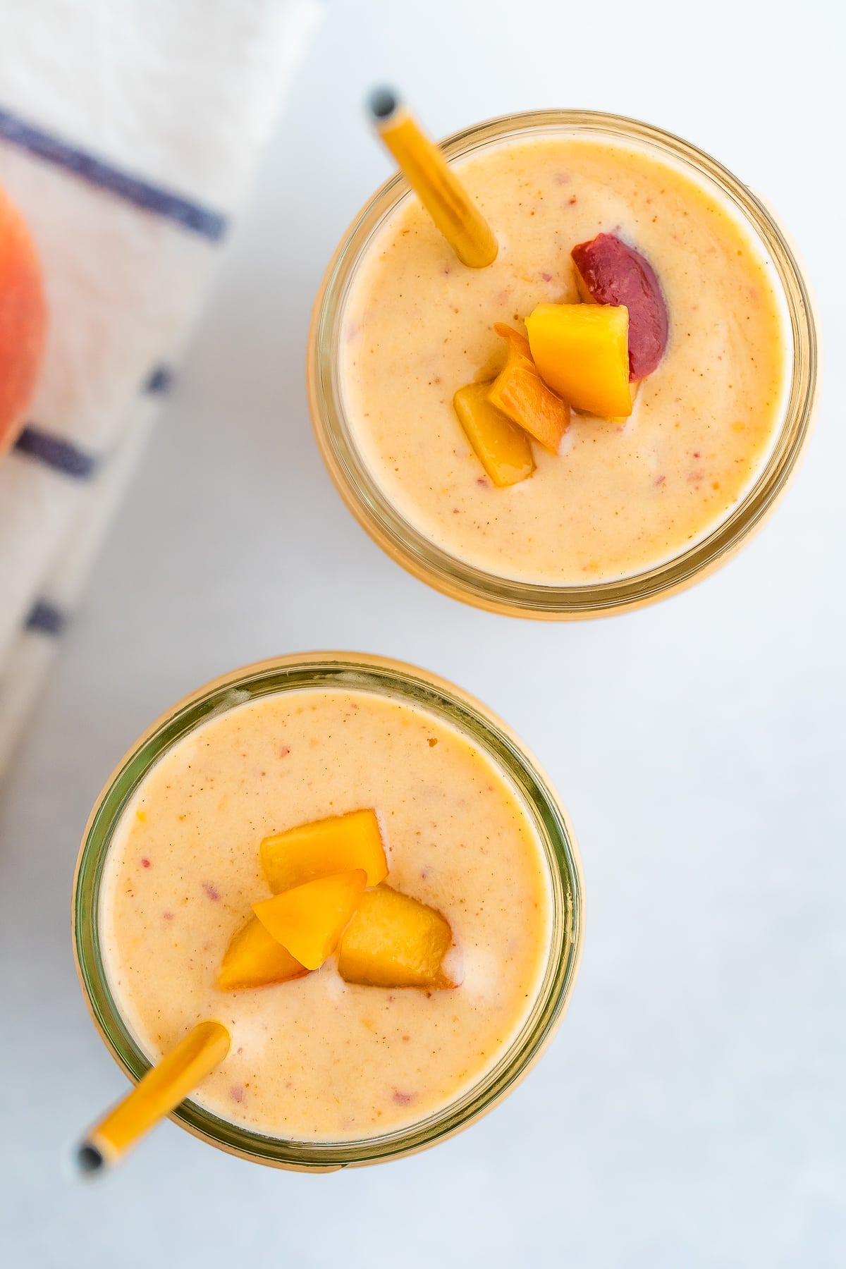 Two glass mason jars of peach smoothies. Two gold straws are in the smoothie and the smoothies are topped with chopped peached.