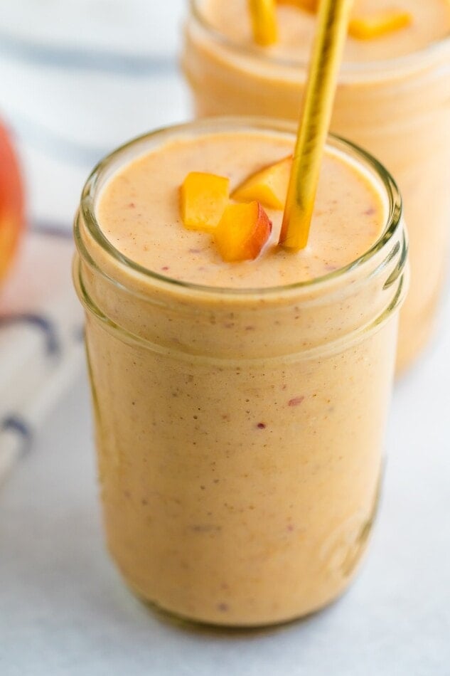 Mason jar with a peach smoothie garnished with a gold straw and chopped peaches.