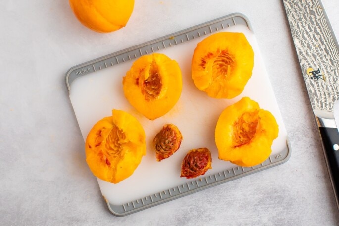 Two peaches on a cutting board with the pits removed and a knife on the right side.