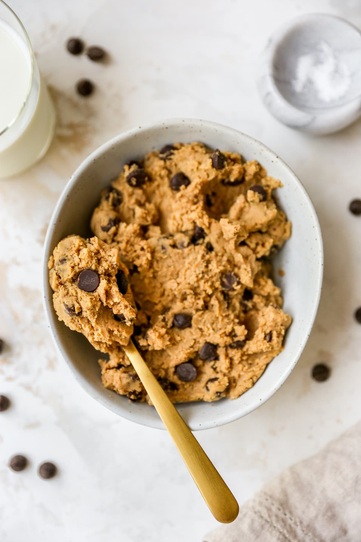 Edible protein cookie dough in a white speckled bowl with chocolate chips on top and gold spoon on the left.