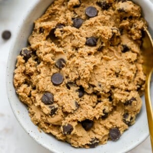 Edible protein cookie dough in a white speckled bowl with chocolate chips on top and gold spoon on the right.