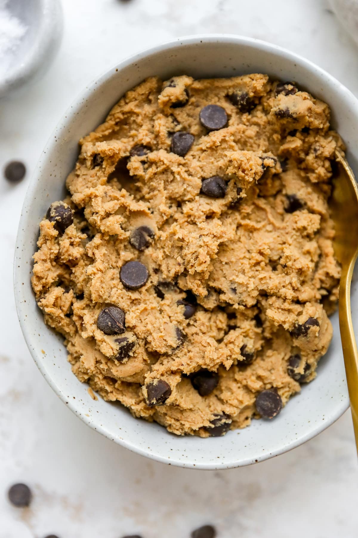 Edible protein cookie dough in a white speckled bowl with chocolate chips on top and gold spoon on the right.