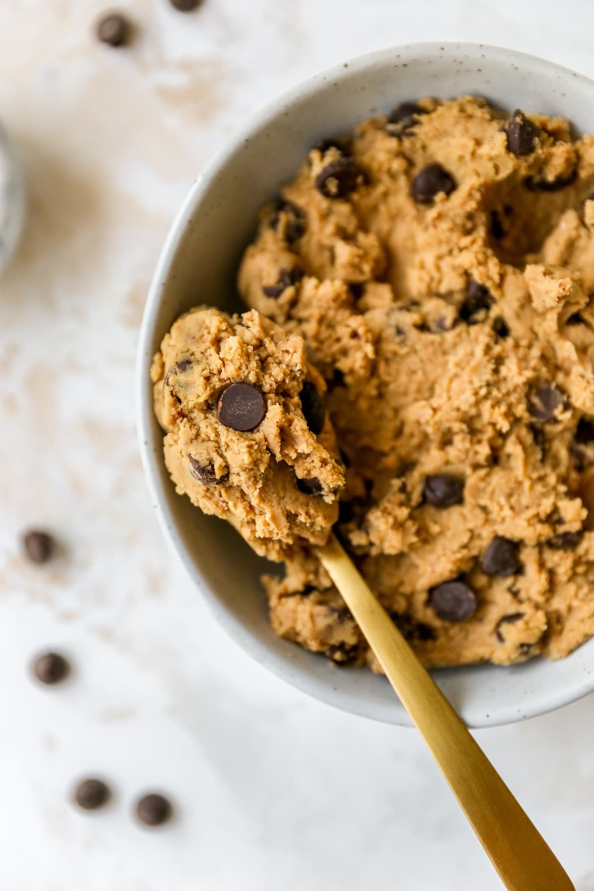 Edible protein cookie dough in a white speckled bowl with chocolate chips on top and gold spoon on the right.