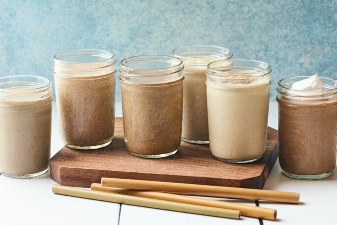 6 protein shakes in Mason jars on a wood board. Bamboo straws are on the table.
