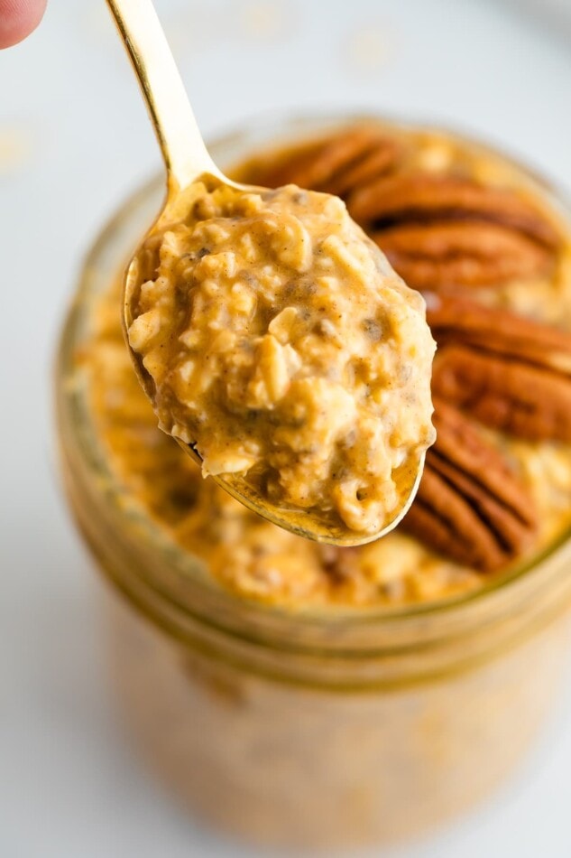 Jar of pumpkin pie overnight oats topped with pecans. Spoon is taking a bite of the oats.