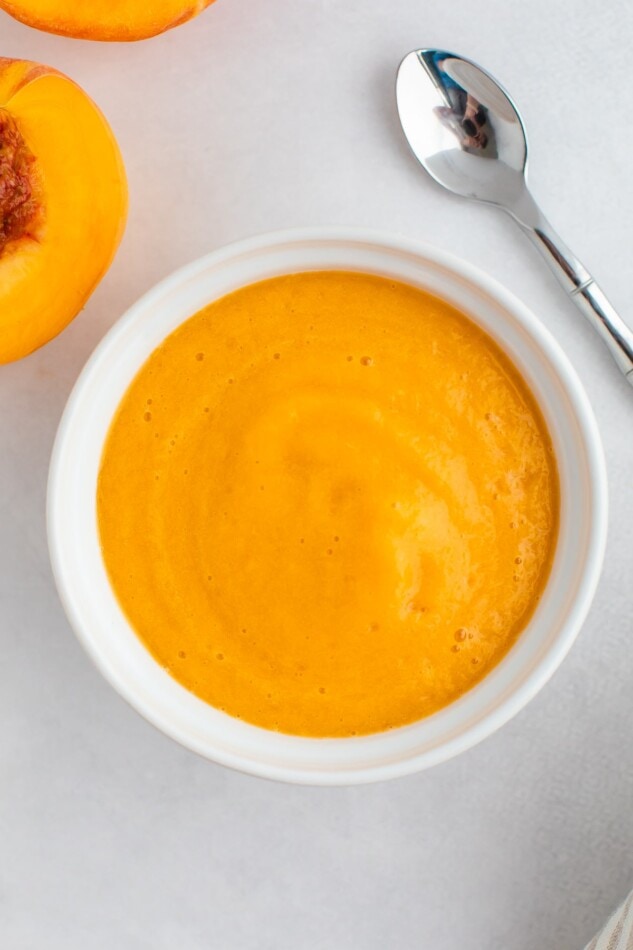 Pureed peaches in a white bowl on a marble surface with a small silver spoon on the side.