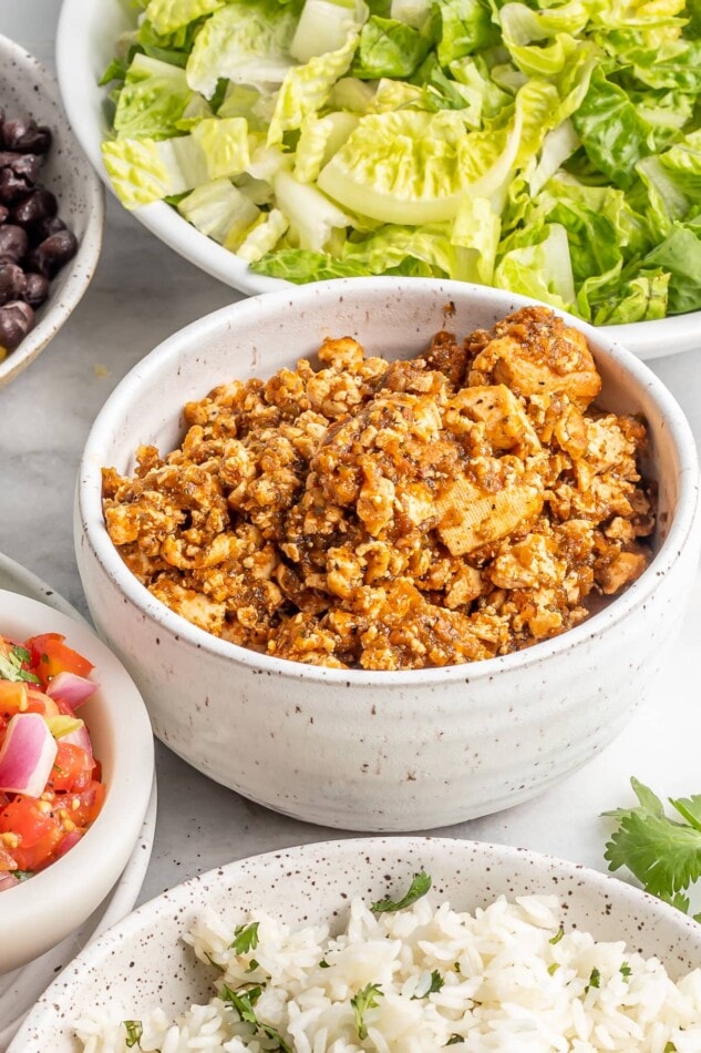 Close up photo of a bowl of sofritas. Other bowls are around it with beans, salsa, lettuce and rice.