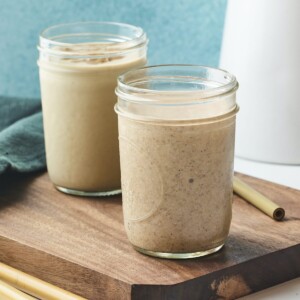 Two vanilla protein shakes in two mason jars. Jars are on a wood cutting board surrounded by bamboo straws.