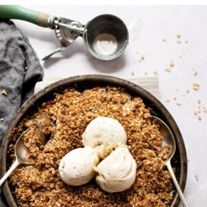 Apple crisp in a pan topped with three scoops of ice cream. A couple spoons are in the pan and an ice cream scoop is beside the pan.