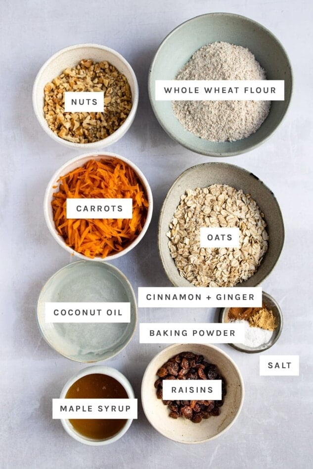 Ingredients measured out to make carrot cake breakfast cookies: nuts, wheat flour, shredded carrots, oats, coconut oil, cinnamon, ginger, baking powder, salt, maple syrup and raisins.