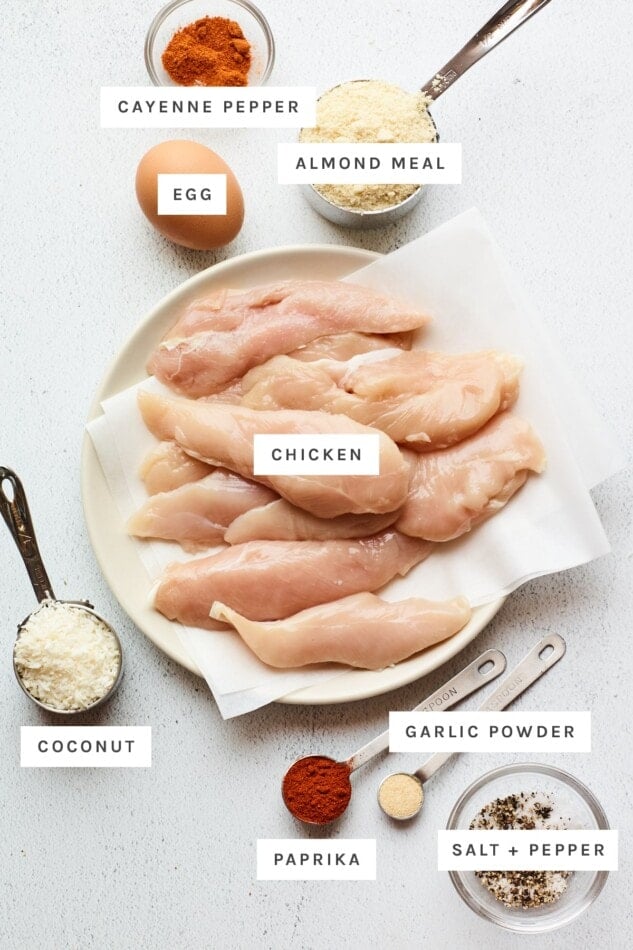 Ingredients measured out to make baked chicken tenders: cayenne pepper, almond meal, egg, chicken, coconut flakes, garlic powder, paprika, salt and pepper.