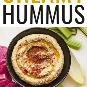 Creamy hummus with olive and spices on top in a black serving bowl with celery and chopped veggies displayed around the bowl.