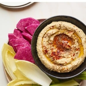 Creamy hummus with olive and spices on top in a black serving bowl with sliced vegetables for dipping plated around the bowl.