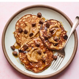 Two plates of protein pancakes topped with chocolate chips, maple syrup and peanut butter.