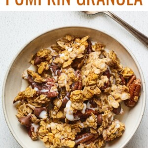 A bowl of pumpkin granola served in a bowl with milk. A silver spoon rests aside the bowl.