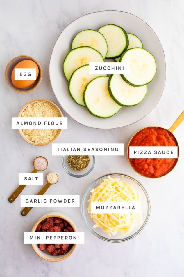 Ingredients measured out to make zucchini pizza bites: egg, zucchini, almond flour, Italian seasoning, pizza sauce, salt, garlic powder, mozzarella and a pepperoni stick.