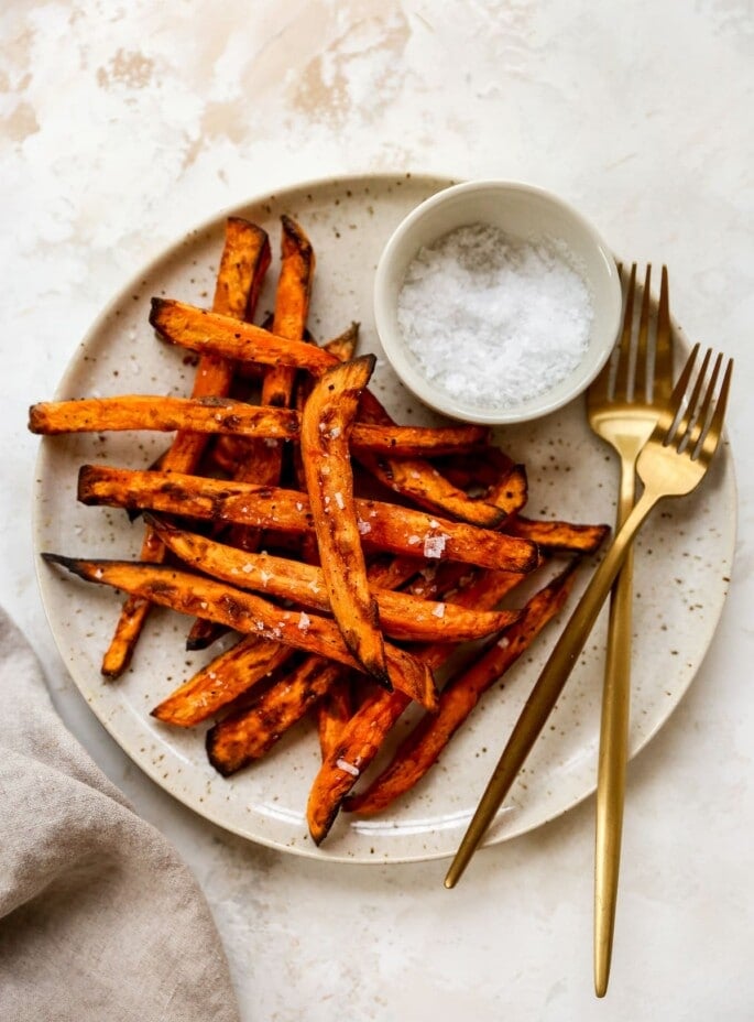 A plate of air fryer sweet potato fries with two forks and a ramekin of salt.