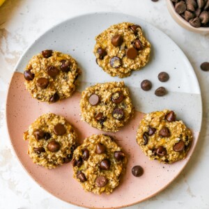 An overhead shot of 6 cookies on a plate with chocolate chips sprinkled around. You can also see a banana and cup of chocolate chips in the top corners.