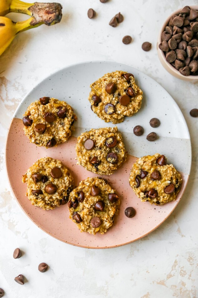 An overhead shot of 6 cookies on a plate with chocolate chips sprinkled around. You can also see a banana and cup of chocolate chips in the top corners.