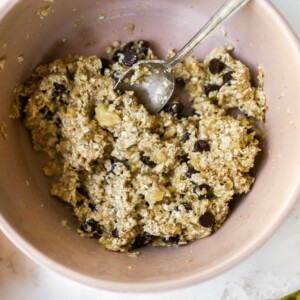 A mixing bowl with the ingredients for banana cookies all mixed together. There is a silver spoon resting inside the bowl.