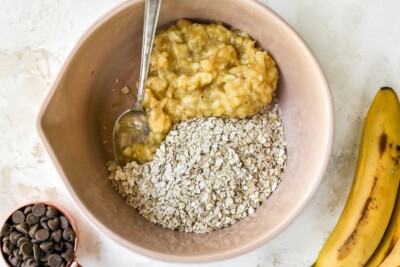 A mixing bowl with oats on one side and mashed banana on the other, prior to being mixed together. A silver spoon rests inside the bowl.