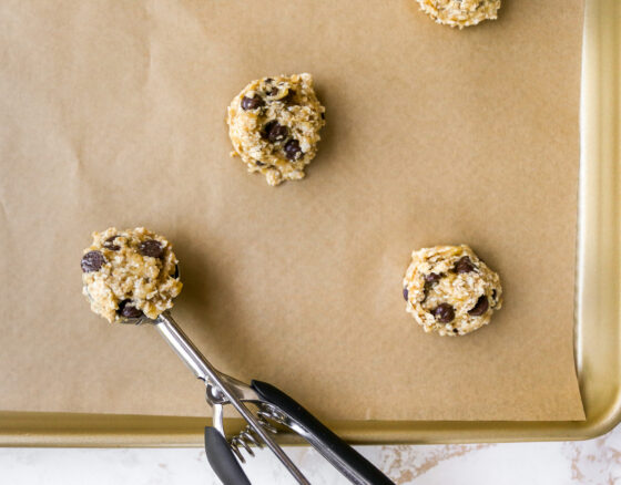 A sheet pan lined with parchment paper with cookies scooped onto it. There is a cookie scoop resting on the sheet pan.