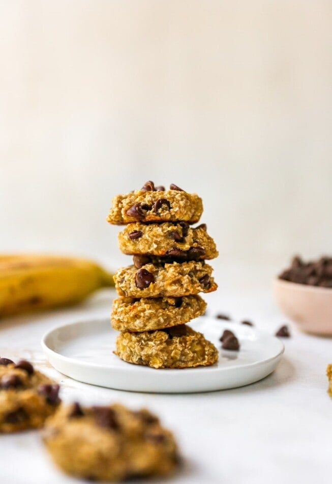 A stack of 5 banana cookies on a white plate, there are two cookies in the bottom left out of focus and ingredients in the background.