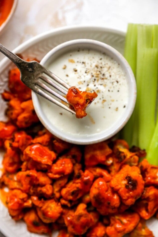 Plate of buffalo cauliflower wings served with ranch and celery sticks. A fork has a cauliflower wing and is dipping it in the ranch.