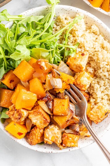 Overhead view of butternut squash tempeh bake served with brown rice and greens, a fork is scooping a bite.