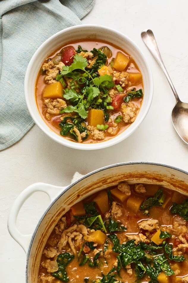 A bowl of butternut squash turkey chili topped with cilantro. A serving pot of chili is cut halfway out of frame.