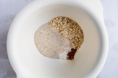 A bowl containing oats, flour, baking powder, cinnamon, salt, and ginger, ready to be combined.