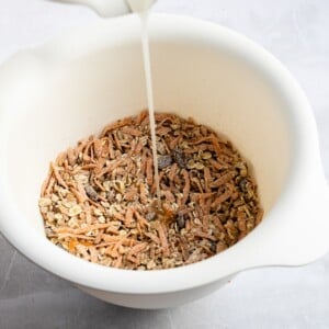 A bowl with a stream of maple syrup being added to a dry mixture of oats, carrots, raisins, pecans, and spices.