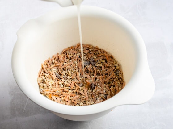 A bowl with a stream of maple syrup being added to a dry mixture of oats, carrots, raisins, pecans, and spices.