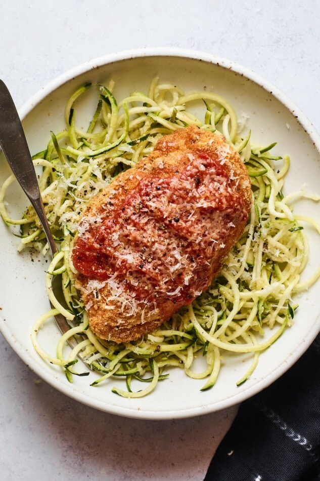 An overhead shot of a bowl zucchini noodles with a chicken parm breast resting on top. A fork rests inside the bowl.