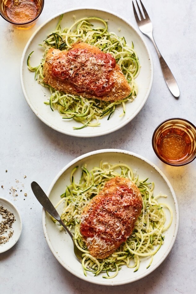 Overhead shot of a healthy dinner dish with chicken parmesan and broccoli. Both topped with a little cheese. An overhead shot of two bowls of zucchini noodles with a chicken breast resting on top of each.