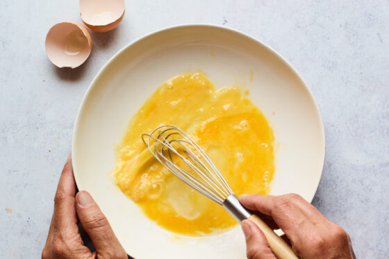 A hand is holding a bowl steady while the other hand is whisking an egg.