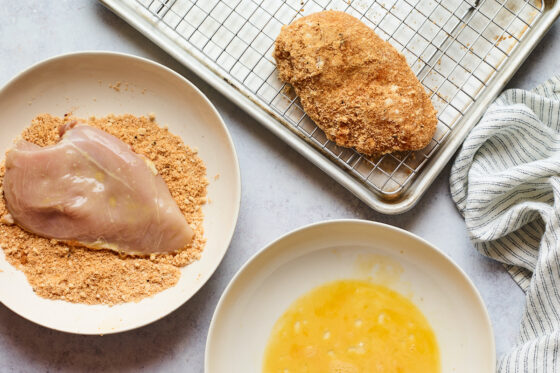 A chicken breast rests in a bowl, being coated with breading, while another chicken breast rests on a metal sheet pan. There is an egg wash bowl in the frame.