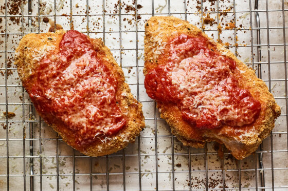 Two breaded chicken breasts covered with marinara sauce and cheese sprinkled on top. They rest on a metal baking sheet.