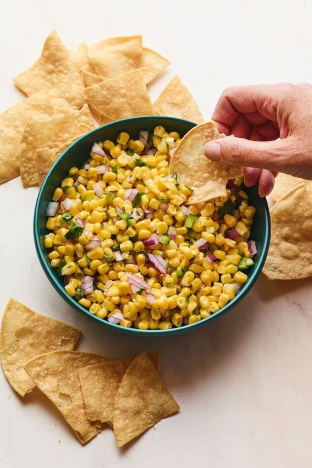 Hand dipping tortilla chip into bowl of easy corn salsa served in blue bowl.