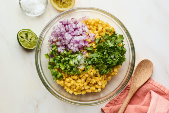 White corn, jalapeño, cilantro, red onion, and lime juice in a glass mixing bowl, chopped and ready to be stirred together.