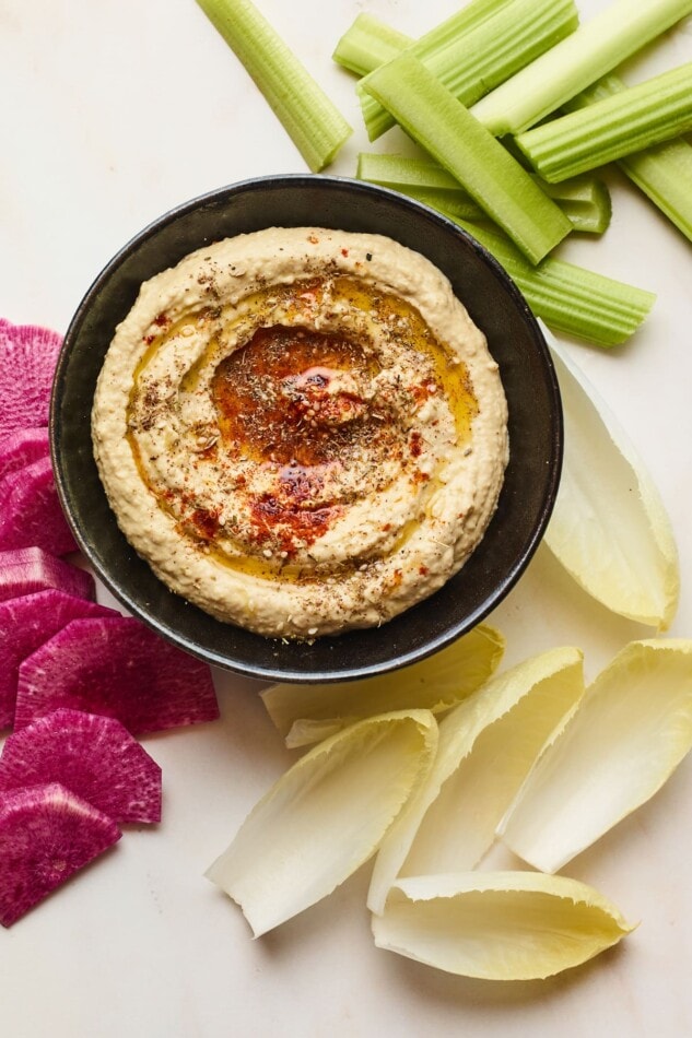 Creamy hummus with olive and spices on top in a black serving bowl with celery and chopped veggies displayed around the bowl.