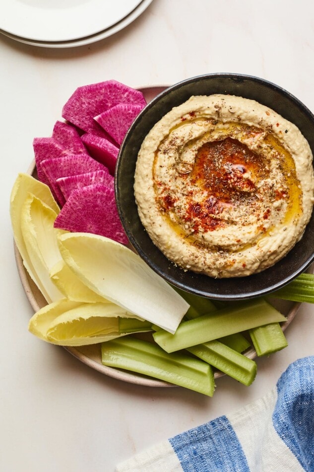 Creamy hummus with olive and spices on top in a black serving bowl with sliced vegetables for dipping plated around the bowl.