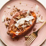 A pink plate with a dessert sweet potato on top. Topping ingredients are sprinkled around the dish and the tabletop. A gold fork rests on the plate.