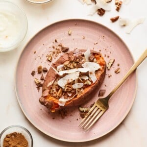 A pink plate with a dessert sweet potato on top. Topping ingredients are sprinkled around the dish and the tabletop. A gold fork rests on the plate.