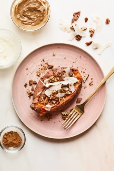 A pink plate with a dessert sweet potato on top. Topping ingredients are sprinkled around the dish and the tabletop. A gold fork rests on the plate.