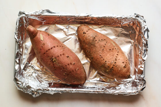 A small baking sheet covered in tin foil with two sweet potatoes on top. The sweet potatoes have holes poked throughout them.