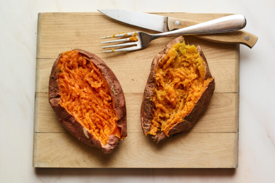 A wooden cutting board with two baked sweet potatoes on top. The sweet potatoes have been sliced open with the insides mashed. There is a fork and knife resting on the cutting board.