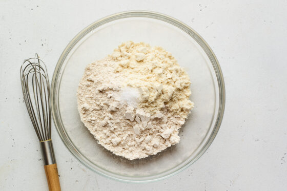 Oat flour and vanilla protein powder in a bowl. Whisk is beside the bowl.